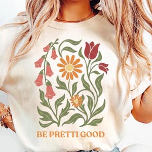 May include: Cream-colored t-shirt featuring a floral design with orange, red, and green elements. The design includes a large daisy, bell-shaped flowers, and the text "BE PRETTI GOOD" in orange letters.