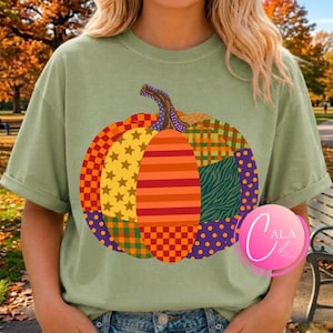 May include: A sage green short-sleeve t-shirt featuring a large, central pumpkin graphic. The pumpkin is composed of various colorful patchwork segments, including patterns of orange stripes, yellow stars, purple dots, green plaid, and zebra print.