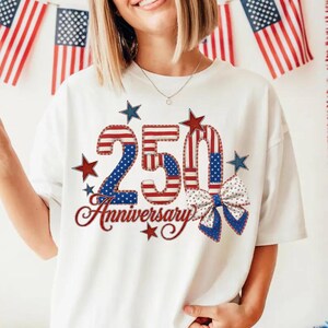 May include: White t-shirt with a patriotic design celebrating a 250th anniversary. The design features the number "250" in red, white, and blue, along with stars and a bow. The word "Anniversary" is written in a cursive font.