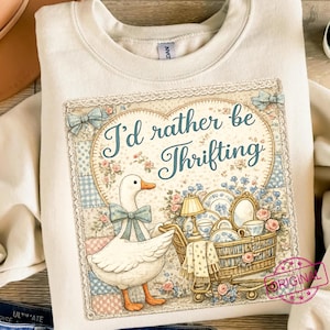 May include: Cream-colored sweatshirt featuring a graphic with a heart-shaped design and the text "I'd rather be Thrifting." The graphic includes a white goose with a blue bow, pushing a shopping cart filled with vintage items.