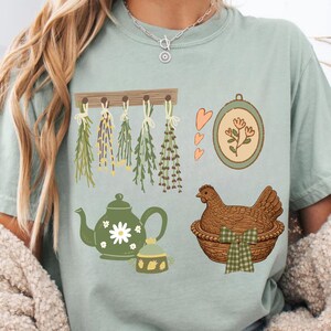 May include: A light green t-shirt featuring various illustrations. Designs include hanging herbs, a teapot with a daisy, a framed floral design, a small jar, a hen in a basket, and three hearts. The shirt has a relaxed fit.