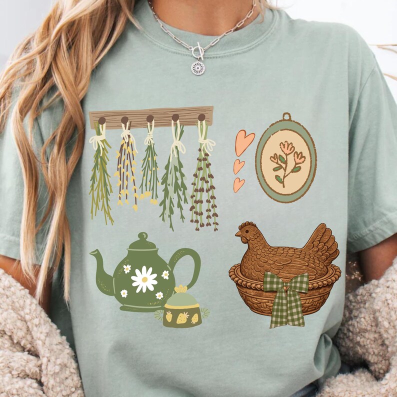 May include: A light green t-shirt featuring various illustrations. Designs include hanging herbs, a teapot with a daisy, a framed floral design, a small jar, a hen in a basket, and three hearts. The shirt has a relaxed fit.