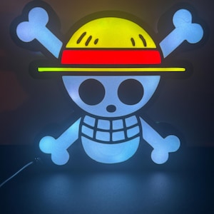 Puede incluir: Un letrero LED brillante con el símbolo de Jolly Roger del anime One Piece. El diseño incluye una calavera blanca con huesos cruzados, un sombrero de paja amarillo con una banda roja y un contorno negro. El letrero emite una luz azul y amarilla.