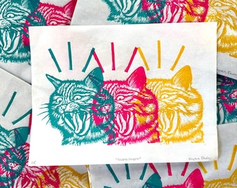 May include: A white paper print featuring three overlapping cat heads in teal, magenta, and yellow. Each cat has an open mouth showing teeth and whiskers. The text "Triata Mophe" and "Kayden Farrier" is handwritten in pencil at the bottom.