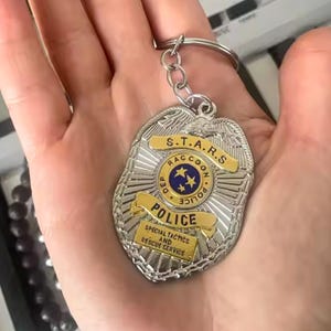 Op de afbeelding: Een zilverkleurige sleutelhanger met een politiebadge-ontwerp en gouden accenten. De badge heeft de tekst "S.T.A.R.S. Raccoon Police" en "Special Tactics and Rescue Service". De sleutelhanger wordt in een hand gehouden.