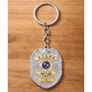 Op de afbeelding: Zilverkleurige metalen sleutelhanger met een politiebadge-ontwerp. De badge heeft de tekst "S.T.A.R.S. Raccoon Police Dept." en "Special Tactics and Rescue Service." De sleutelhanger heeft een ketting en een ronde sleutelring.