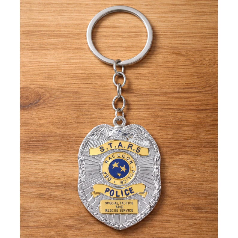 Op de afbeelding: Zilverkleurige metalen sleutelhanger met een politiebadge-ontwerp. De badge heeft de tekst "S.T.A.R.S. Raccoon Police Dept." en "Special Tactics and Rescue Service." De sleutelhanger heeft een ketting en een ronde sleutelring.
