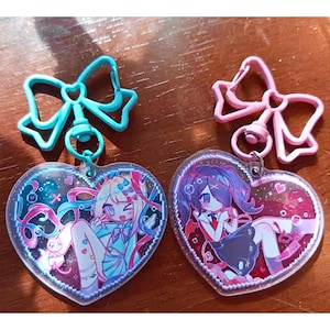 May include: Two heart-shaped keyrings with anime-style illustrations. One has a blue bow-shaped clasp and features a light blue and pink character. The other has a pink bow clasp and a purple and red character.