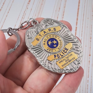 Op de afbeelding: Een zilverkleurige politiebadge sleutelhanger met gouden accenten. De badge toont de tekst "S.T.A.R.S. Raccoon Police Dept." en "Special Tactics and Rescue Service". De sleutelhanger is bevestigd aan een zilveren ring.