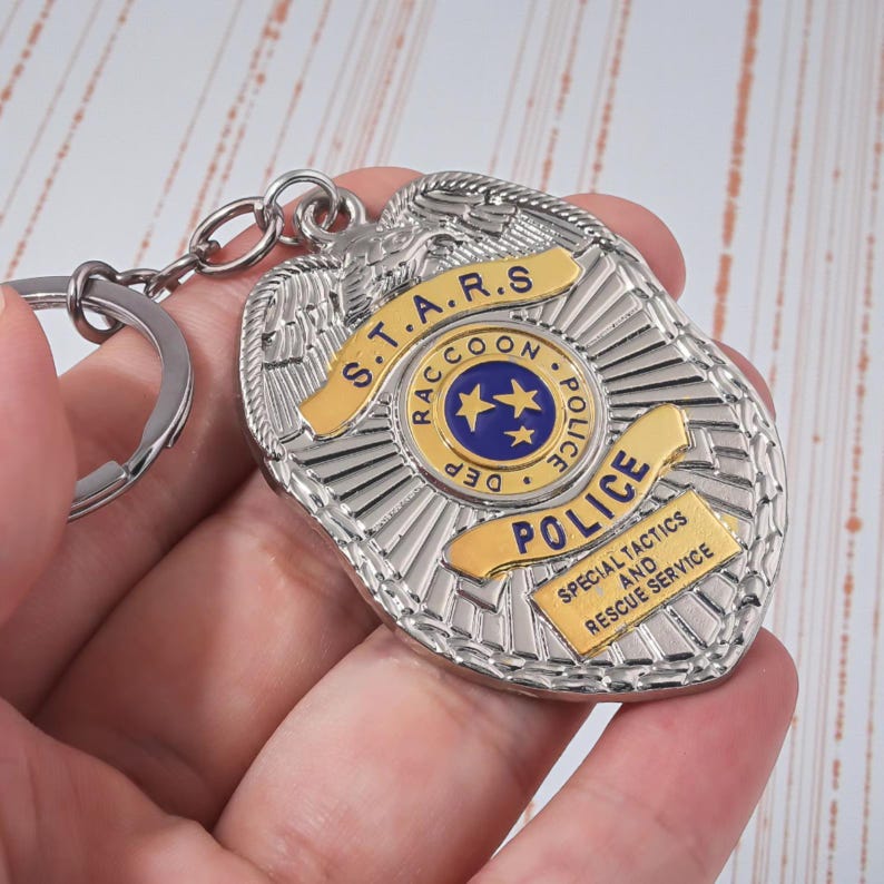 Op de afbeelding: Een zilverkleurige politiebadge sleutelhanger met gouden accenten. De badge toont de tekst "S.T.A.R.S. Raccoon Police Dept." en "Special Tactics and Rescue Service". De sleutelhanger is bevestigd aan een zilveren ring.