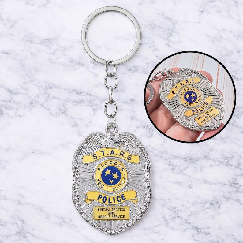 Op de afbeelding: Zilverkleurige metalen sleutelhanger met een politiebadge-ontwerp. De badge heeft een gele banner met de tekst "POLICE" en "S.T.A.R.S. RACCOON DEP". De badge bevat ook de tekst "SPECIAL TACTICS AND RESCUE SERVICE".