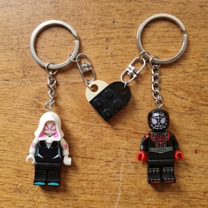Peut inclure: Deux porte-clés avec des figurines attachées à un charme en forme de cœur divisé. L'un a un personnage à capuche blanche avec une combinaison noire, l'autre un personnage en costume noir avec un emblème d'araignée rouge. Le cœur est bicolore.