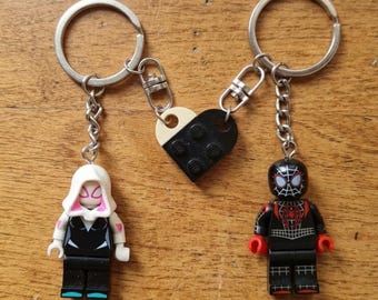 Puede incluir: Dos llaveros con minifiguras unidas a un dije de corazón dividido. Uno tiene un personaje con capucha blanca y traje negro, y el otro un personaje con traje negro y emblema de araña rojo. El corazón es de color blanco y negro.