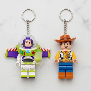 Peut inclure: Deux porte-cl&eacute;s avec des figurines de style Lego. L'un est Buzz l'&Eacute;clair dans une combinaison spatiale blanche, verte et violette avec un casque transparent. L'autre est Woody, un cow-boy avec un chapeau marron, une chemise jaune et un pantalon bleu. Les deux ont des porte-cl&eacute;s argent&eacute;s.