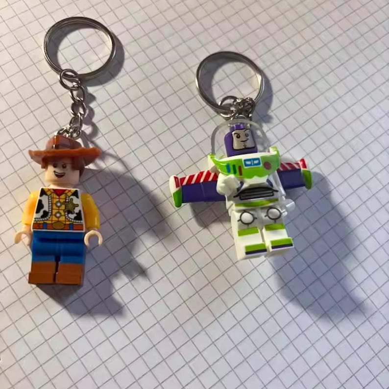 Peut inclure: Deux porte-cl&eacute;s avec des figurines de personnages de style Lego. L'un est un cow-boy avec un chapeau marron, une chemise jaune et un pantalon bleu. L'autre est Buzz l'&Eacute;clair dans une combinaison spatiale blanche et verte avec des accents violets et des ailes.