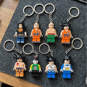 Puede incluir: Una colección de ocho llaveros de personajes con minifiguras de plástico con diversos atuendos y peinados. Cada figura está unida a una anilla de metal plateado mediante una cadena corta, mostrando expresiones pintadas detalladas y ropa colorida.