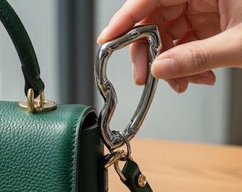 Puede incluir: Un mosquetón de metal plateado sujeto a un bolso de mano de cuero verde oscuro. El mosquetón tiene un diseño curvo único y está sujeto por una mano. El bolso de mano presenta herrajes dorados.