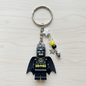 May include: A Batman-themed keychain featuring a black Lego figure with a yellow and black emblem, a black cape, and a silver keyring. The keychain includes a chain with star and heart charms, and yellow, black, and clear beads.