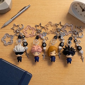 Puede incluir: Cuatro llaveros de personajes de anime con figuras chibi de cabello blanco, rosa, castaño y negro. Cada llavero incluye un cierre en forma de estrella, cuentas blancas y negras, dados, dijes de bola ocho y pequeños detalles de estrellas de metal.