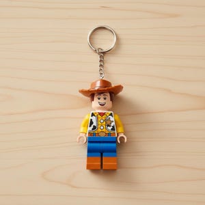 Peut inclure: Un porte-cl&eacute;s de style Lego repr&eacute;sentant un personnage de cow-boy avec un chapeau marron, une chemise jaune, un pantalon bleu et des bottes marron. Le porte-cl&eacute;s a un anneau et une cha&icirc;ne argent&eacute;s. Le personnage a un badge de sh&eacute;rif et un visage souriant.