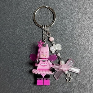 May include: A pink Batman-themed keyring featuring a Lego-style figure with fairy wings, a tutu, and a star-topped wand. The keyring includes a silver ring, decorative bows, pearls, and a star charm. The figure has a pink bat symbol on its chest.
