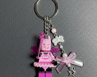 May include: A pink Batman-themed keychain featuring a Lego-style figure with fairy wings, a tutu, and a star-topped wand. The keychain includes a silver ring, decorative bows, pearls, and a star charm. The figure has a pink bat symbol on its chest.