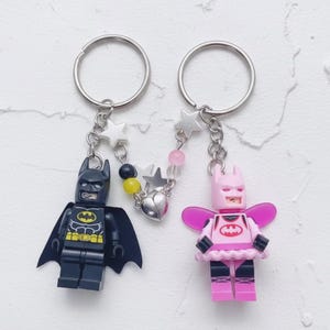 May include: Two keychains featuring Lego-style Batman figures. One is black with a yellow emblem, the other is pink with a tutu and wings. Both have silver keyrings and star-shaped charms.
