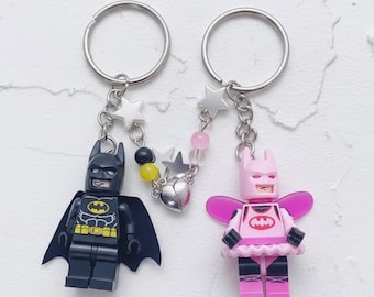 Puede incluir: Dos llaveros con figuras de Batman estilo Lego. Uno es negro con un emblema amarillo, el otro es rosa con tutú y alas. Ambos tienen llaveros plateados y dijes en forma de estrella.