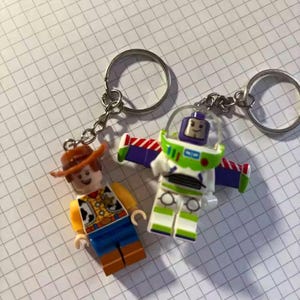 Peut inclure: Deux porte-cl&eacute;s avec des figurines de style Lego de Woody et Buzz Lightyear de Toy Story. Woody porte un chapeau et un gilet marron. Buzz est dans une combinaison spatiale blanche et verte avec des ailes violettes. Les deux ont des porte-cl&eacute;s argent&eacute;s.
