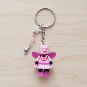May include: A pink Batman-themed keychain featuring a Lego-style figure with a tutu and wings. The figure has a Batman logo on the chest. The keychain includes a silver ring, a star charm, and pink beads. The background is a light wood surface.