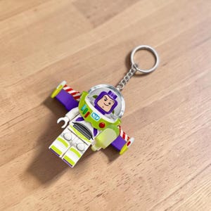 Peut inclure: Un porte-cl&eacute;s Lego Buzz l'&Eacute;clair. Le porte-cl&eacute;s repr&eacute;sente une figurine miniature de Buzz l'&Eacute;clair avec un casque transparent, une combinaison spatiale blanche et verte et des ailes violettes. La figurine est attach&eacute;e &agrave; un porte-cl&eacute;s en m&eacute;tal argent&eacute;.