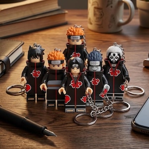 Puede incluir: Una colección de seis llaveros de personajes con figuras estilizadas que visten capas negras con patrones de nubes rojas. Cada figura tiene un peinado y expresión facial únicos, unidos a una cadena de metal plateado y una anilla circular.