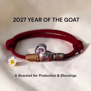 May include: A dark red cord bracelet featuring a silver-toned goat charm with a small coin detail. The charm is flanked by a tan thread wrap and a multicolored thread wrap. Text reads: 2027 YEAR OF THE GOAT and A Bracelet for Protection & Blessings.