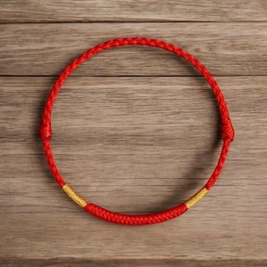 Puede incluir: Una pulsera circular de cordón rojo con una textura trenzada en toda la banda. El diseño incluye dos pequeños acentos cilíndricos de color dorado colocados simétricamente a lo largo de la sección inferior del cordón.