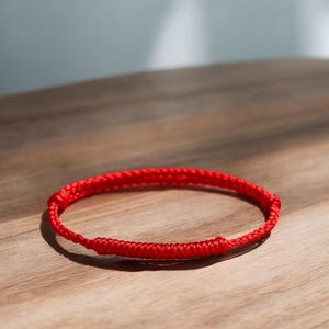 Puede incluir: Una pulsera de cordón rojo vibrante con un diseño trenzado y dos secciones anudadas cilíndricas. La pulsera está construida con una longitud continua de hilo rojo fino y suave, formando una estructura circular con nudos ajustables.
