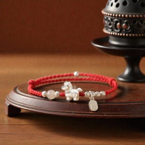Puede incluir: Una pulsera trenzada roja con un dije de caballo plateado, una cuenta redonda blanca y pequeñas cuentas plateadas. Un pequeño dije plateado con caracteres chinos cuelga de la pulsera. La pulsera se exhibe sobre una superficie de madera.