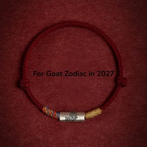 May include: A red cord bracelet featuring a central silver-toned cylindrical bead engraved with a goat motif. The bracelet includes decorative multi-coloured thread accents on either side of the bead and the text For Goat Zodiac in 2027.