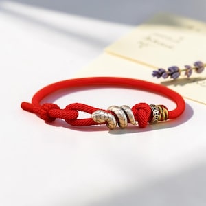 May include: A red cord bracelet featuring a metallic snake-shaped charm and a small cylindrical bead with engraved patterns. The bracelet is secured with a knotted closure, showcasing a vibrant red colour and detailed metal accents on the cord.