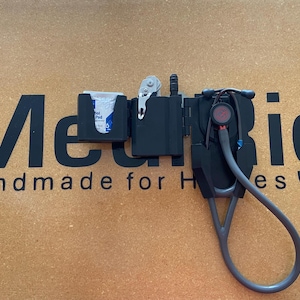 May include: A black medical organizer holster featuring a dedicated slot for a stethoscope, a small pocket containing a white alcohol prep pad, and a central compartment holding a silver trauma shear and a black penlight, all arranged horizontally.