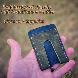 May include: A handcrafted olive and sapphire leather cardholder. The wallet is made from full-grain Italian leather and features white stitching along the edges. The text "Badalassi Carlo Pueblo Full Grain Italian Leather Olive and Sapphire" is visible.