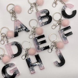 Può includere: Portachiavi con alfabeto con un design in resina nera e rosa, scaglie d'argento e un soffice pompon rosa. Ogni lettera ha un portachiavi e una chiusura argentati. Le lettere A, B, C, D, E, F, G, H e J sono visibili.