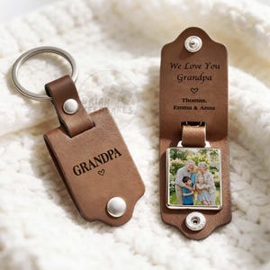 May include: Brown leather keychain with the word "GRANDPA" and a heart engraved on it. The keychain is attached to a silver metal ring. Another keychain has a photo of a family and the text "We Love You Grandpa" with names.