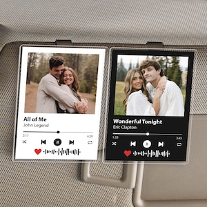 May include: Two clear acrylic car visor clips with photos of couples. One features "All of Me" by John Legend, the other "Wonderful Tonight" by Eric Clapton. Each clip has a play button, song length, and a heart icon.