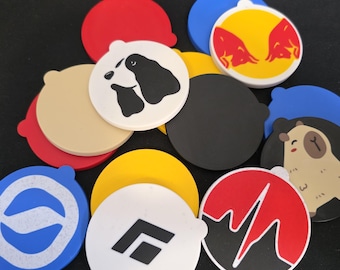 May include: A collection of colorful, round silicone coasters. Designs include a dog and puppy, a capybara, a logo with a lightning bolt, and a logo with a stylized letter F. Other coasters are solid red, black, yellow, and blue.