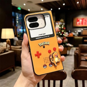 Könnte beinhalten: Gelbe Handyhülle im Design eines Game Boy Advance SP. Mit dem Pokémon-Logo, einem roten Kreuz, roten Knöpfen und einer Charmander-Figur. Die Hülle hat einen schwarzen Rand und einen weißen Bereich für die Kamera.