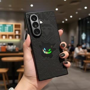 Peut inclure: Coque de téléphone noire avec un motif texturé et un personnage de dessin animé lumineux. L'étui a une découpe pour un appareil photo à trois objectifs et un motif en relief détaillé. Le personnage a des accents verts et bleus. L'étui est tenu dans une main avec du vernis à ongles noir.