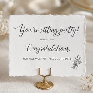 Peut inclure: Une carte rectangulaire blanche aux bords effilochés maintenue par un petit support en métal doré. La carte présente un texte gris foncé : You're sitting pretty! Congratulations. YOU HAVE WON THIS TABLE'S CENTERPIECE! Une illustration florale.