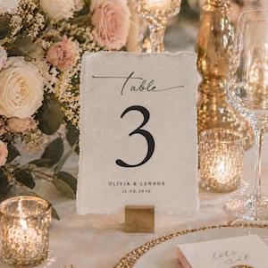 Peut inclure: Mise en place de mariage avec composition florale, bougies et carton de numéro de table. Le carton indique « Table 3 » avec les noms Olivia & Lennox et la date 11.02.2030. Le cadre comprend des accents dorés et de la verrerie.