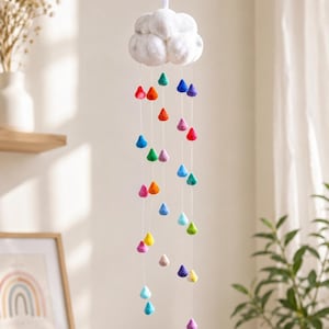 May include: A white cloud-shaped mobile with colorful felt raindrops. The raindrops are in various colors, including red, orange, yellow, green, blue, and purple. The mobile hangs from a white ribbon, suitable for a nursery or children's room.