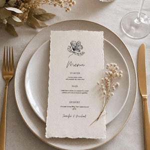 Może przedstawiać: Elegancka aranżacja stołu weselnego z kartą menu, talerzem i złotymi sztućcami. Karta menu zawiera ilustrację kwiatową i słowo "Menu". Menu zawiera pozycje przystawek, dań głównych i deserów. Suszone kwiaty i świeca.
